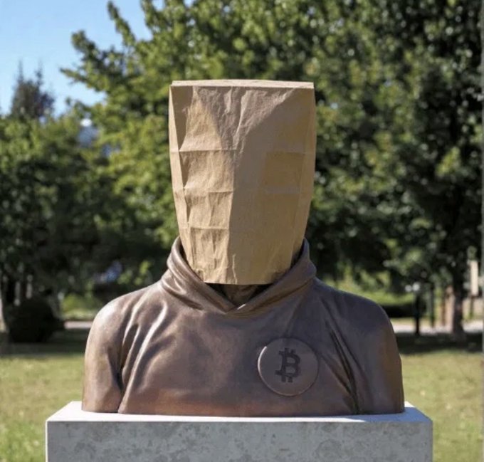 A bronze statue of a hooded figure wearing a paper bag over its head with a Bitcoin symbol on the chest, displayed outdoors on a pedestal.