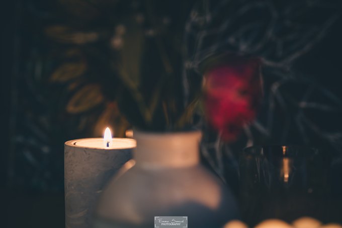 The image features a white vase with a red flower sitting on top of a table next to two candles, one of which has its flame burning brightly while the other's light is dimmed. The background appears dark, but there are some leaves visible in the scene. The focus of the image is on the vase and flowers, creating an atmosphere of tranquility and calmness.