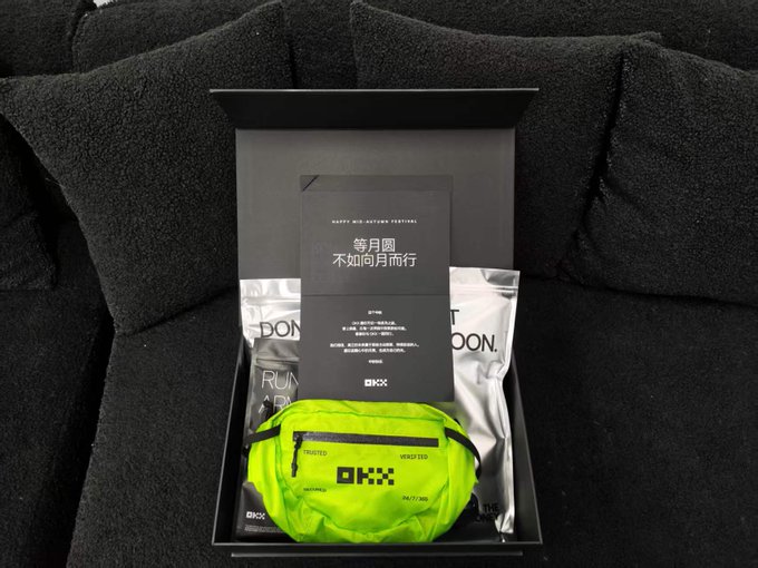 An open black gift box on a black couch containing a neon green running belt labeled 'OKX' and other branded items with a card featuring Chinese text and 'Happy Mid Autumn Festival'.