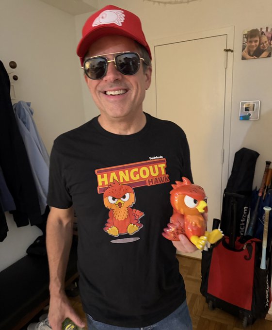 A smiling man wearing a red cap and aviator sunglasses, dressed in a black 'HANGOUT HAWK' t-shirt featuring Angry Birds artwork, holding a matching Angry Birds toy figure in his hand.
