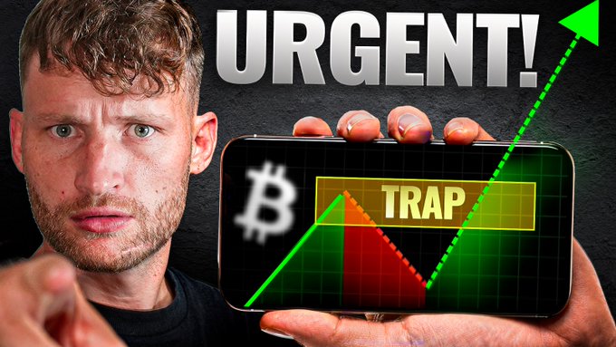 The caption could be:

A man with a serious expression holds a phone displaying a Bitcoin chart with a green upward arrow and a yellow "TRAP" label, while bold "URGENT!" text appears above, illustrating the post's warning about a temporary pullback before a larger pump in the crypto market.