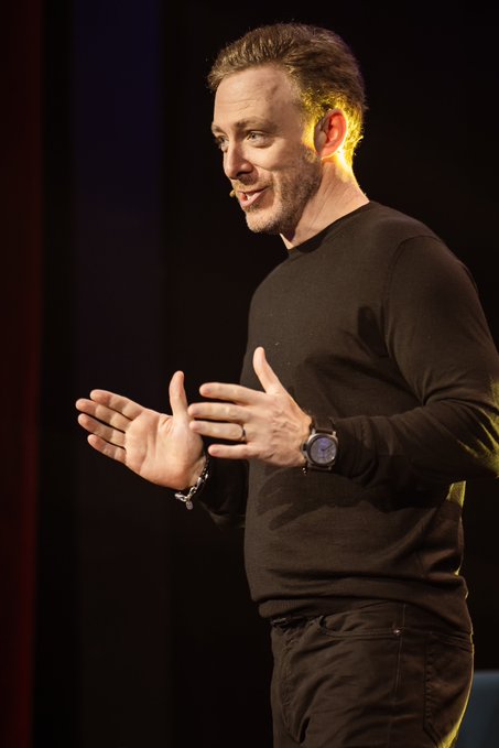 A man with short blond hair, wearing a black long-sleeve shirt and a wristwatch, stands on a dimly lit stage gesturing expressively with both hands as he speaks to an audience. The image captures him mid-sentence, fitting the context of a speaker engaging in a discussion or Q&A about blockchain technology, specifically posing a question about which blockchain is technically superior to ICP.