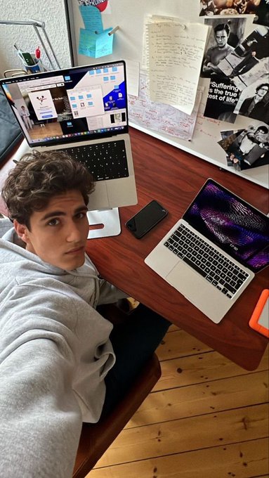 A young man in a grey hoodie sitting at a wooden desk with two laptops and a smartphone, with various papers and posters on the wall behind him.