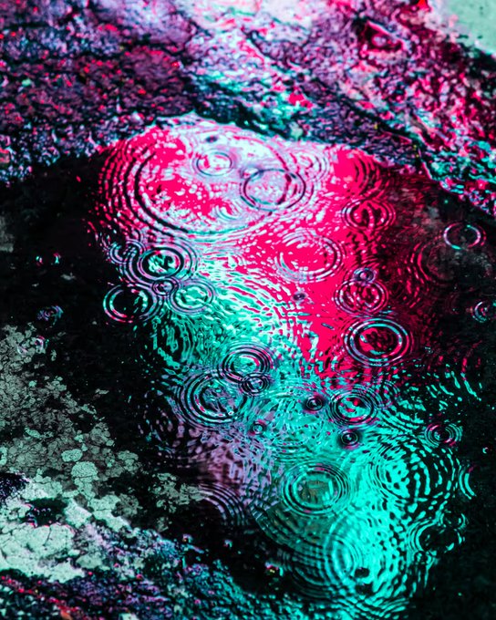 A vibrant, close-up image of raindrops creating concentric ripples in a puddle, illuminated with striking magenta and teal hues that give it an almost otherworldly appearance. The dreamlike, psychedelic colors beautifully complement the poetic "drip-drop" theme, evoking the surreal and artistic style associated with Dave Krugman's photography.