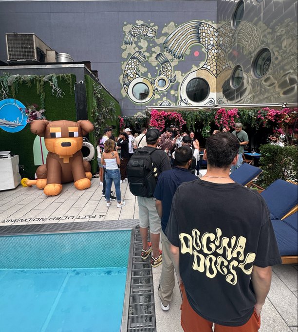 A crowd gathers at an outdoor venue featuring a large inflatable bear figure, colorful koi fish wall art, and a man wearing a black 'DIGNA DOGG' t-shirt standing by a swimming pool.