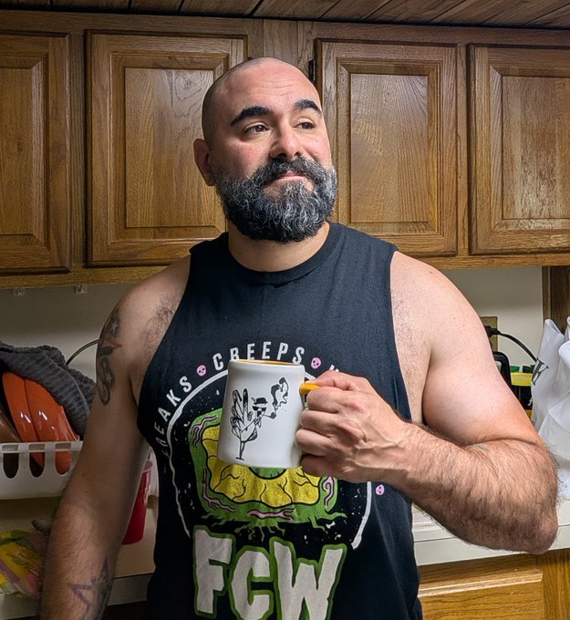 A bearded, bald man in a black "Freaks Creeps FCW" tank top holds a white mug with a creepy tree character design, standing in a kitchen before wooden cabinets, fitting the post's teaser about an upcoming Non Fungible Creeps announcement.