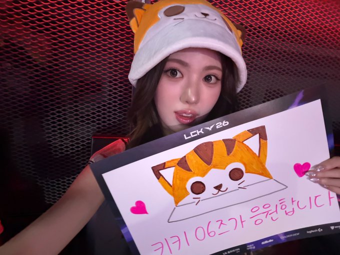 A young woman wearing an orange and white cat-themed hat holds a sign featuring a hand-drawn orange cat and Korean text that reads "카키 여즈가 응원합니다" (Kaki Girls cheering), with "LCK Y26" visible at the top.