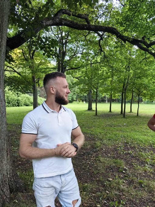 A bearded man wearing a white polo shirt and ripped jeans poses outdoors under the shade of a large tree in a park.