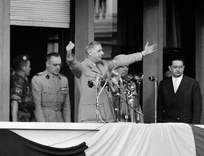 The image features a man standing at a podium with his arms raised, possibly giving a speech or addressing an audience. Behind him, there are several microphones set up for use by the people present. The background includes other individuals who appear to be listening intently as well.
