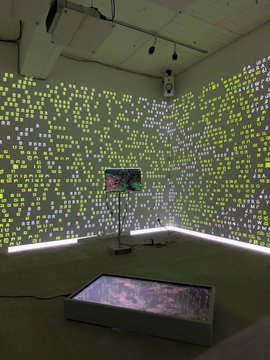 A dimly lit art installation room where yellow-green pixelated text and symbols are projected across the white walls, with a small screen on a stand and a glowing rectangular display on the floor.