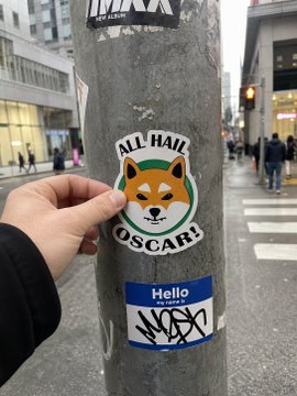 A hand applying a circular sticker that reads 'ALL HAIL OSCAR!' featuring a Shiba Inu dog face to a metal pole on a city street, below a 'Hello' sticker.