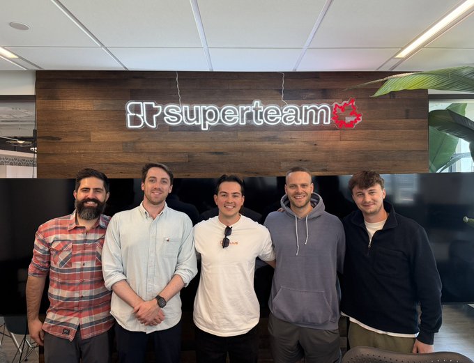In the image, a group of five men are standing together in front of a wooden wall with a neon sign that reads "Superteam" above them. The men appear to be posing for a photo, possibly at an event or gathering related to their company's arrival in Waterloo, as indicated by the post.