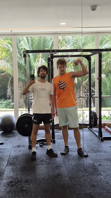 Two men posing for a photo in a home gym with a squat rack, one holding a phone for the mirror selfie.