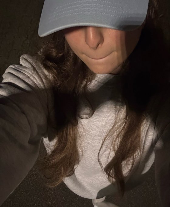 A close-up, high-angle selfie shows a person with long brown hair wearing a light blue baseball cap and a white t-shirt, with the cap obscuring their eyes.