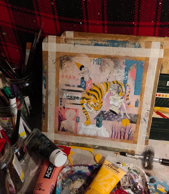 A mixed-media collage artwork on a wooden board depicting a tiger and a woman in a surreal scene, surrounded by paint tubes and brushes on a work surface.