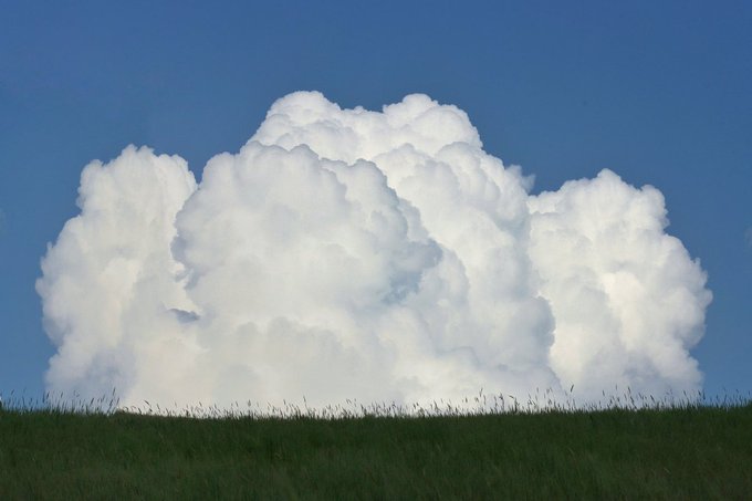 A large, billowing white cumulus cloud rises above a green grassy hill against a clear blue sky.