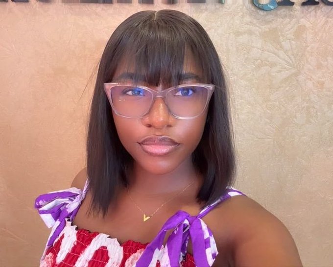 A close-up selfie of a woman with dark hair, bangs, and clear-framed glasses with a blue tint, wearing a colorful floral top and a small gold triangle necklace.