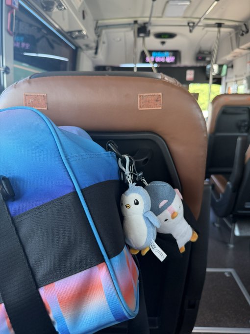 Inside a bus, a blue and black backpack hangs on a seat back with two small plush keychains—a blue penguin and a white bird with a pink bow.