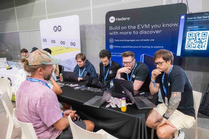 A team of hackers collaborates around laptops at the Hedera booth during ETHGlobal Lisbon, working on autonomous agent projects aimed at winning a share of the $15,000 prize pool. The booth signage reads "Hedera - Build on the EVM you know, with more to discover," highlighting the platform's native capabilities and AI tooling for developers.