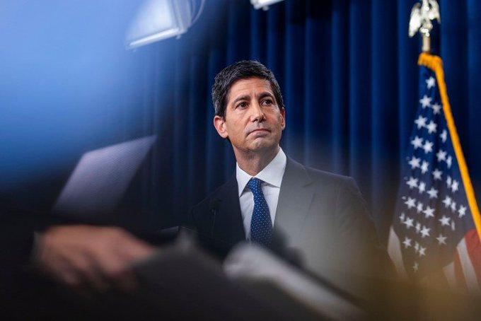 A man in a dark suit and blue tie stands in front of a U.S. flag and blue curtain, appearing to be at a formal event or press conference.