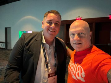 A selfie of two men, one in a gray blazer wearing a lanyard and the other in an orange 'PopSockets' t-shirt, posing together indoors at what appears to be a conference or event.
