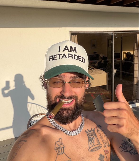 A bearded man with tattoos, sunglasses, and a large diamond chain smiles and gives a thumbs-up while smoking a cigar, wearing a white trucker hat with bold black text reading "I AM RETARDED." The self-deprecating humor of the hat pairs with the "Day 1 of selfie a day until SOL hits ATH" post to convey an irreverent, bullish commitment to Solana and to documenting the journey.