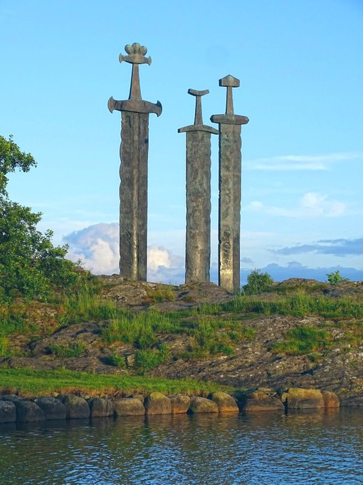 Three large stone swords of different shapes stand upright on a rocky hill beside water, silhouetted against a blue sky.