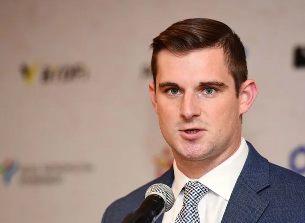 A young man in a navy suit and checkered tie speaks into a microphone at what appears to be a press event, with a blurred backdrop featuring sponsor logos.