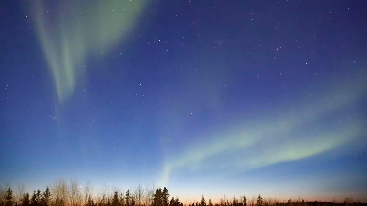 The image captures a breathtaking view of the Aurora Borealis, also known as the Northern Lights, dancing across the night sky. The lights are vibrant green and blue hues that stretch from the top left corner towards the bottom right corner of the frame. The sky is filled with numerous stars, creating an awe-inspiring backdrop for this natural spectacle. The image is taken during the Blue Hour, which is a period just after sunset when the sun is low on the horizon, casting long shadows and bathing everything in a soft, warm light that enhances the colors of the Aurora Borealis.