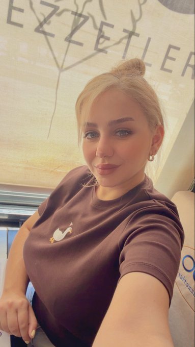A selfie of a blonde woman with her hair in a bun, wearing a brown t-shirt with a small bird emblem, seated indoors with a 'EZKL ETIE' banner behind her.