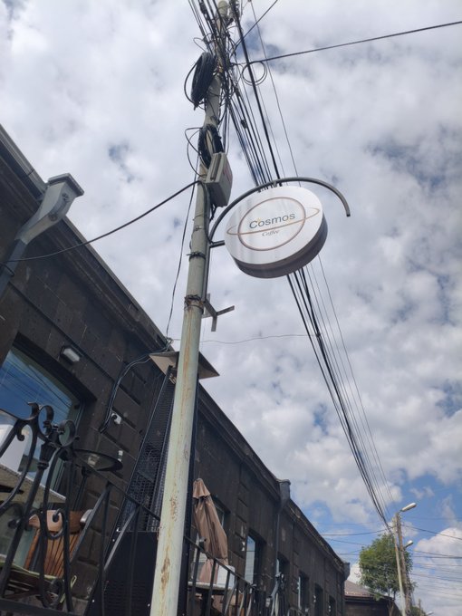 A weathered utility pole displays a round "Cosmos" sign, surrounded by tangled electrical wires against a cloudy sky, capturing the faded commercial presence that makes the caption's quip about Cosmos's dwindling relevance particularly fitting.