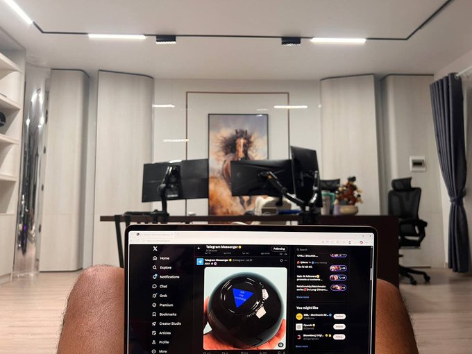 A person holds a laptop showing the Telegram Messenger desktop app with a chat titled 'DALL-E AI' featuring a black sphere with a blue triangle, while in the background sits a modern gaming/streaming setup with dual monitors, an office chair, and a large framed artwork on the wall.