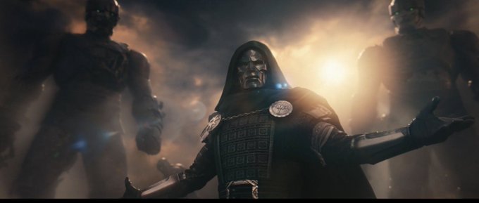 A cinematic still shows a hooded figure in metallic armor with a mask standing in front of two towering stone-like figures, backlit by a bright light source.