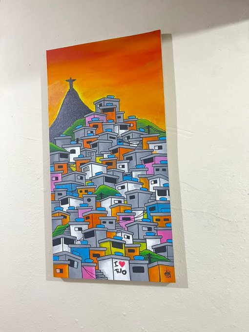 Photograph of a vibrant street-art style painting depicting a colorful favela hillside with Christ the Redeemer statue silhouetted against an orange sunset sky.