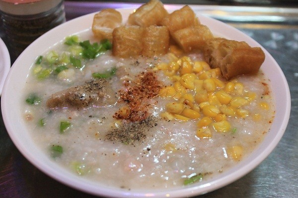 A bowl of Chinese congee or porridge topped with yellow corn kernels, green scallions, fried dough sticks (youtiao), and sprinkled with seasonings.
