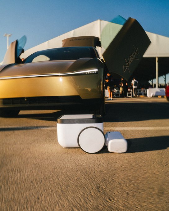 A gold futuristic concept car with its doors open, displayed outdoors at an event with a white and teal building in the background and small white devices in the foreground.