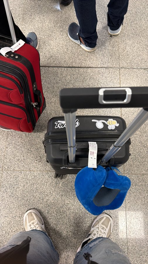 Overhead view of a black suitcase and a red suitcase on a tiled airport floor with two people standing nearby and a blue neck pillow attached to a luggage handle.