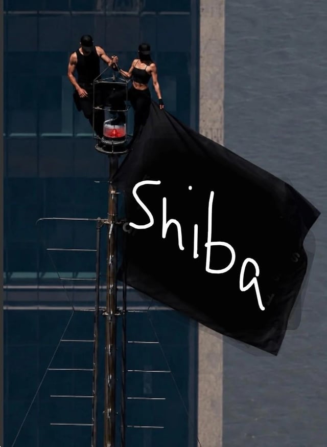 Two performers in black outfits stand on a tall structure, holding a large black flag with 'Shiba' written in white handwritten-style text.
