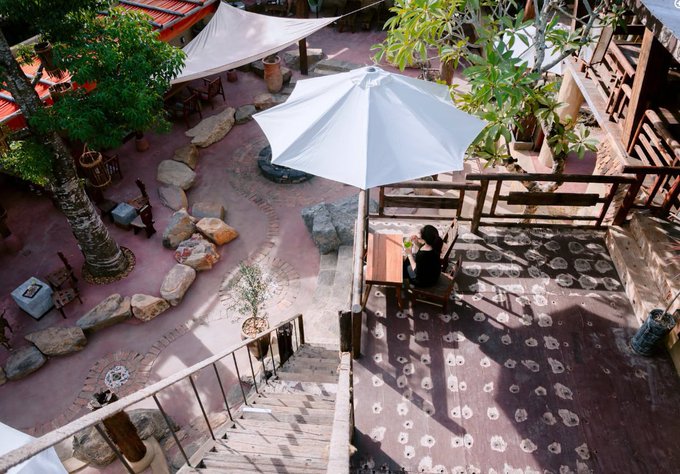 An aerial view of a rustic outdoor courtyard with red brick flooring, stone pathways, a large white umbrella, scattered rocks, plants, and a person seated at a wooden table on a balcony.