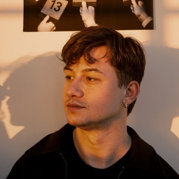 A portrait of a young man with brown hair and a slight mustache, wearing a dark jacket and earring, lit with warm golden light against a wall with artwork behind him.