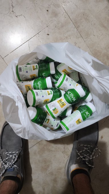 A white plastic bag filled with empty green and white Activia yogurt bottles sits on a tiled floor between a person's gray boat shoes.