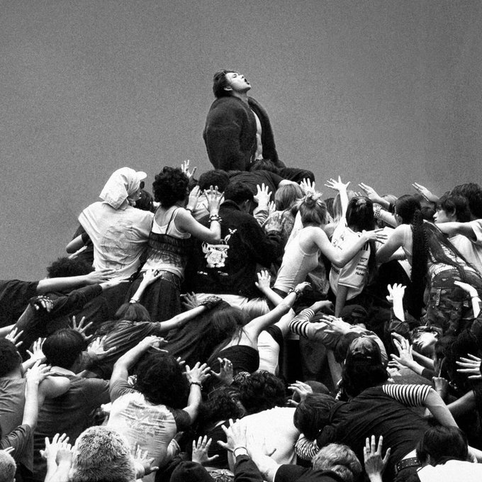 A black and white photograph showing a man in a suit standing on a pile of people with their hands reaching upward, with a 'BTS' watermark in the corner.