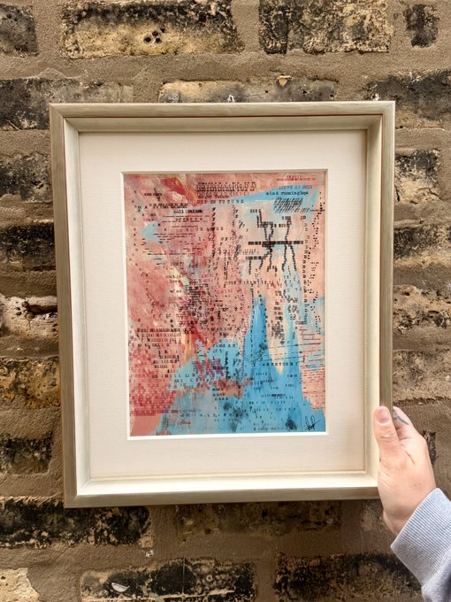 A hand holds up a framed abstract collage artwork featuring pink and blue washes with scribbled text against a rustic brick wall, captioned "fin" (French for "end") to signal the conclusion of the artistic piece.