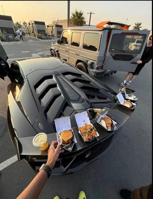 The image shows a luxury car—appearing to be a Lamborghini—and a Mercedes G-Wagon parked in a lot with four fast food meals (burgers and sides in takeout containers) lined up across the hood, evoking a humorous "flex" of wealth paired with casual dining. The accompanying post jokes about OpenSea's potential token launch spanning 2021-2022 activity, playfully contrasting high-value crypto gains with the meme of rich crypto traders flaunting wealth over fast food.