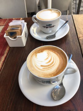 Two cups of cappuccino featuring latte art, served on white saucers with spoons on a wooden table next to a sugar packet holder.