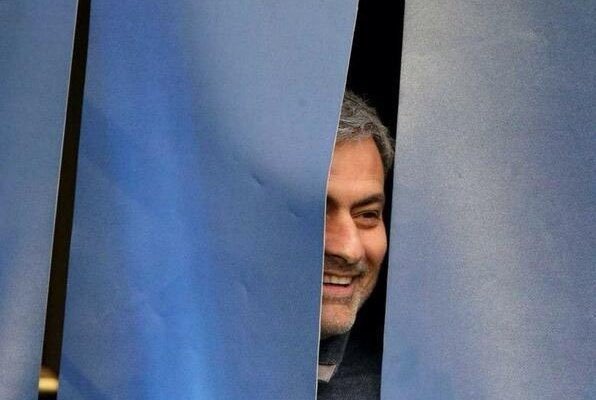 A man with grey hair and a beard smiles warmly as he peeks through a narrow gap in blue curtains.