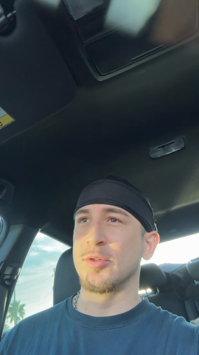 A selfie of a young man wearing a black headband and blue t-shirt, sitting in the driver's seat of a car with a cloudy sky visible through the window.