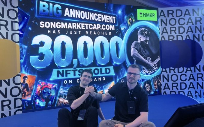 Two men shaking hands in front of a large screen displaying a 'BIG ANNOUNCEMENT: SONGMARKETCAP.COM HAS JUST REACHED 30,000 NFTS SOLD ON CARDANO,' featuring an 'NMKR' logo and an image of a guitarist.
