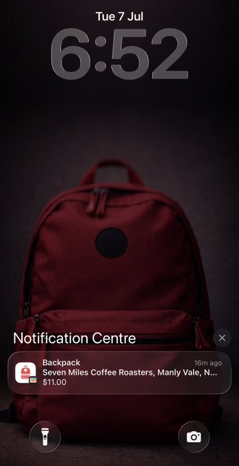 An iPhone lock screen at 6:52 AM on Tuesday, July 7th, showing a maroon backpack as the wallpaper with a Notification Centre alert for an Apple Pay transaction of $11.00 at Seven Miles Coffee Roasters in Manly Vale from 16 minutes ago. The image perfectly captures the start of the user's morning routine, featuring both the coffee purchase and their backpack as they head out.