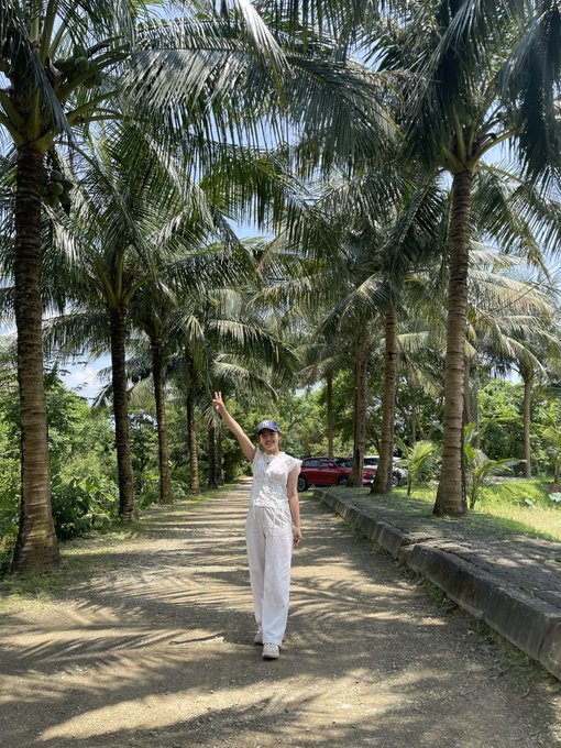 A woman in a white outfit and cap posing with one arm raised in a peace sign on a path lined with tall palm trees, with a red SUV parked nearby.